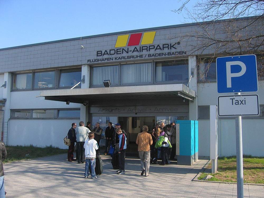 Eingang zum Terminal des Flughafens Karlsruhe/Baden-Baden mit Reisenden und Hinweisschild zu Parkplatz und Taxi